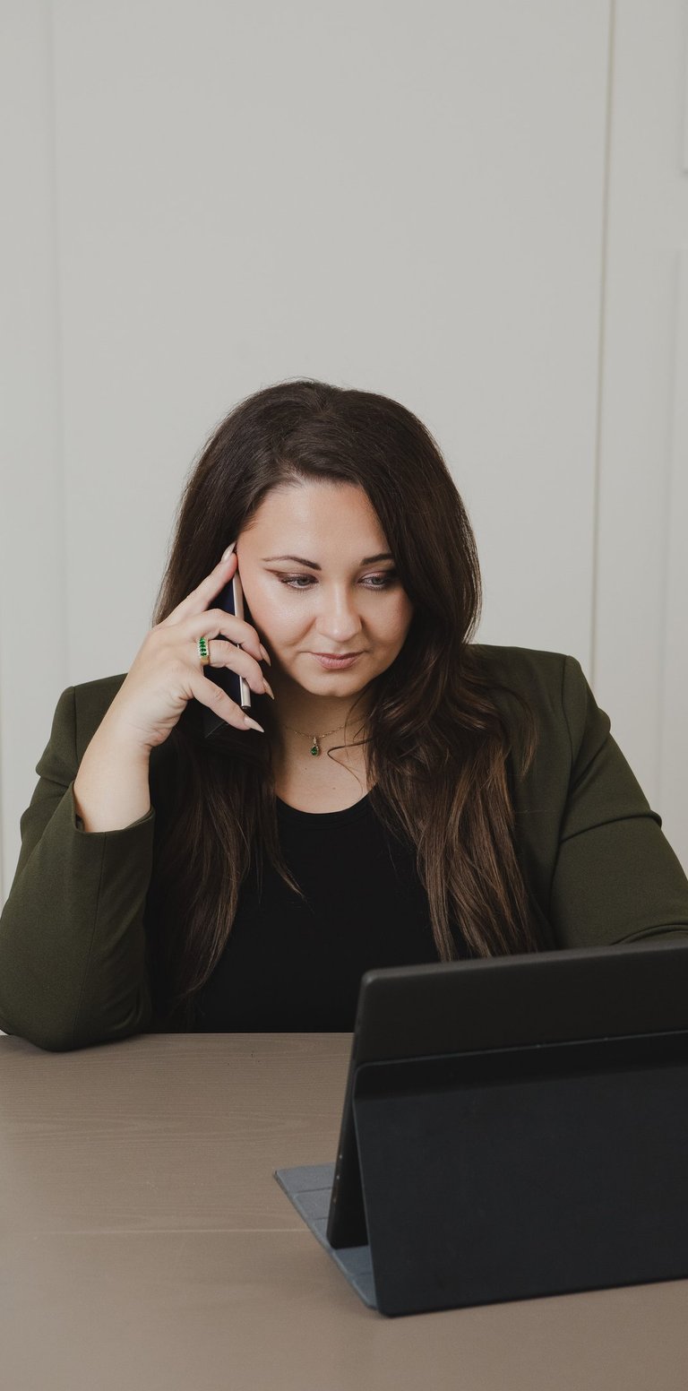 Adriana in olive blazer talking on a smartphone while working on a tablet laptop.