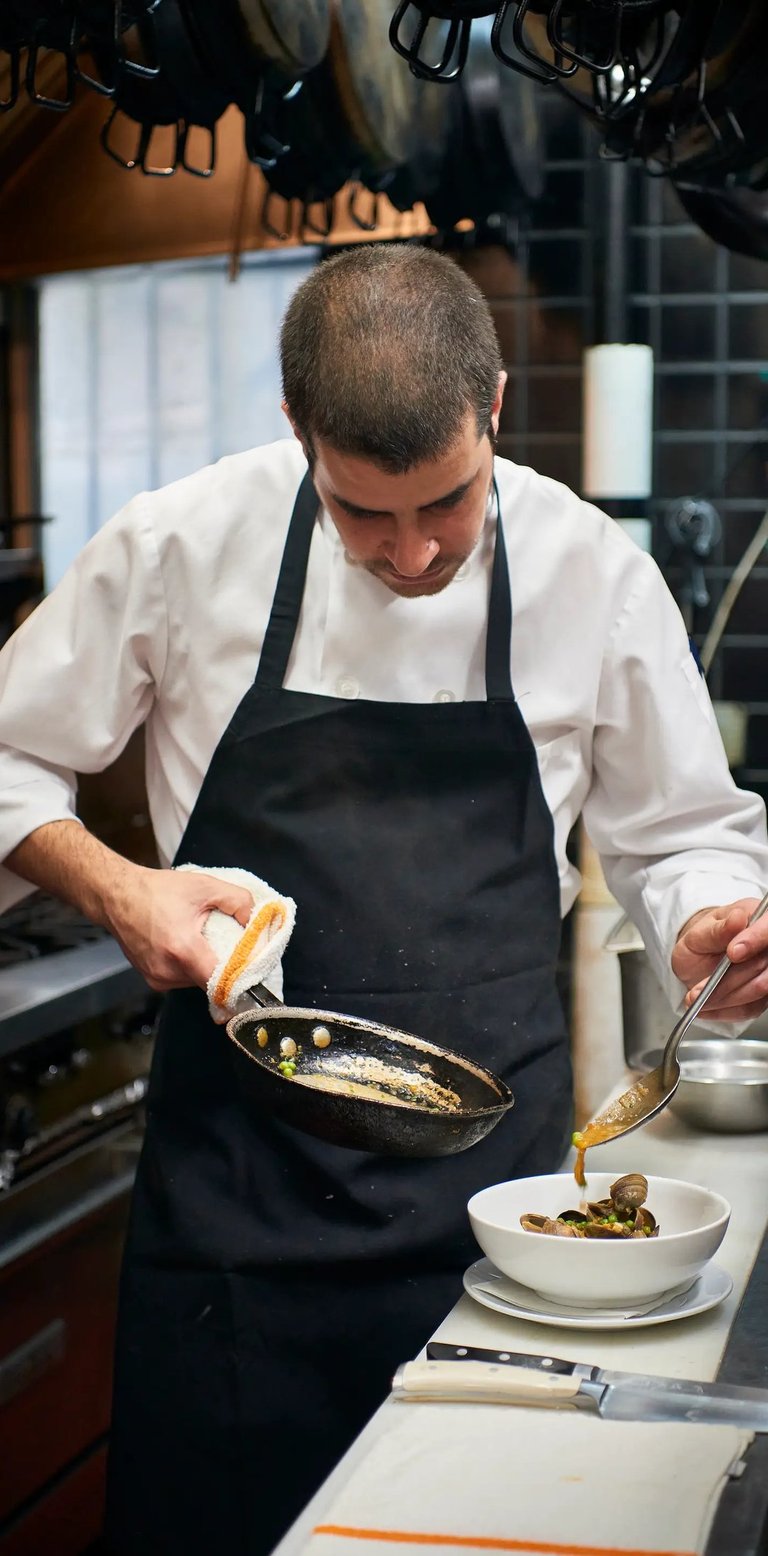 Chef wearing an uniform, pouring sauce from a pan into a bowl of clams in a professional kitchen.