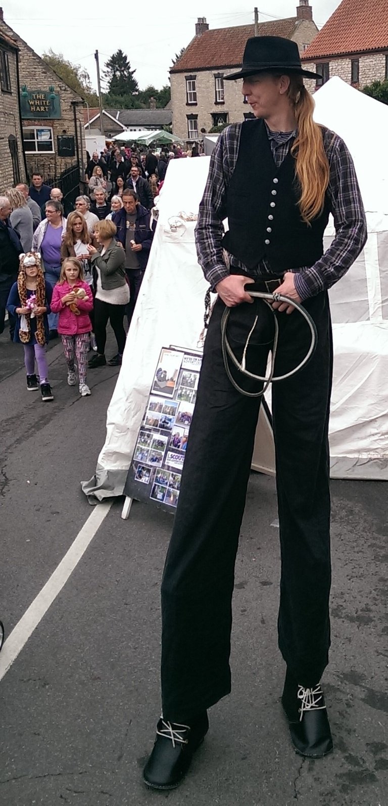 A tall stilt walker performer in a black vest and hat at a crowded outdoor community street festival.
