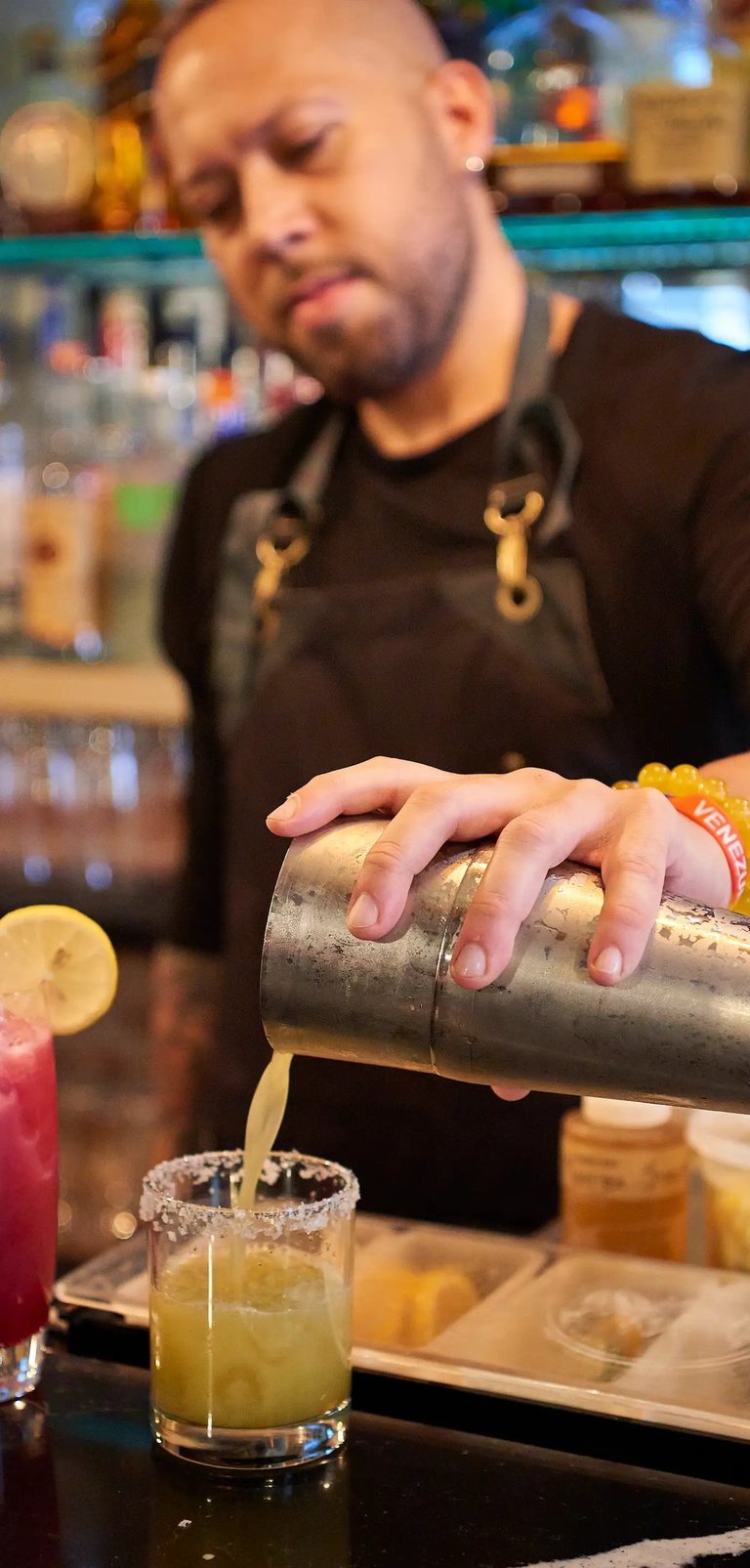Bartender wearing a black apron, pouring a cocktail from a shaker into a glass with a salted rim