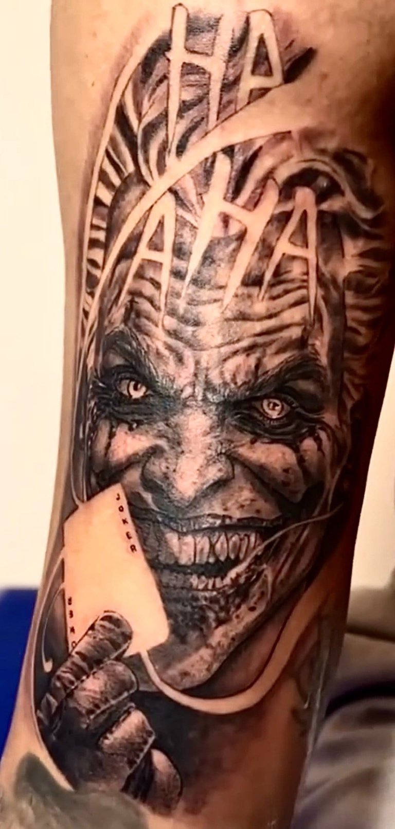 Detailed black and grey Joker tattoo featuring a realistic villainous grin, HA HA text, and a joker playing card.