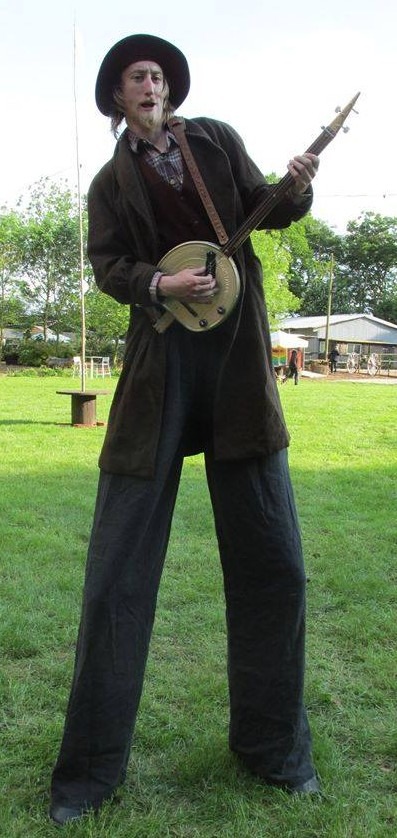A street performer on tall stilts playing a banjo in a grassy park setting.