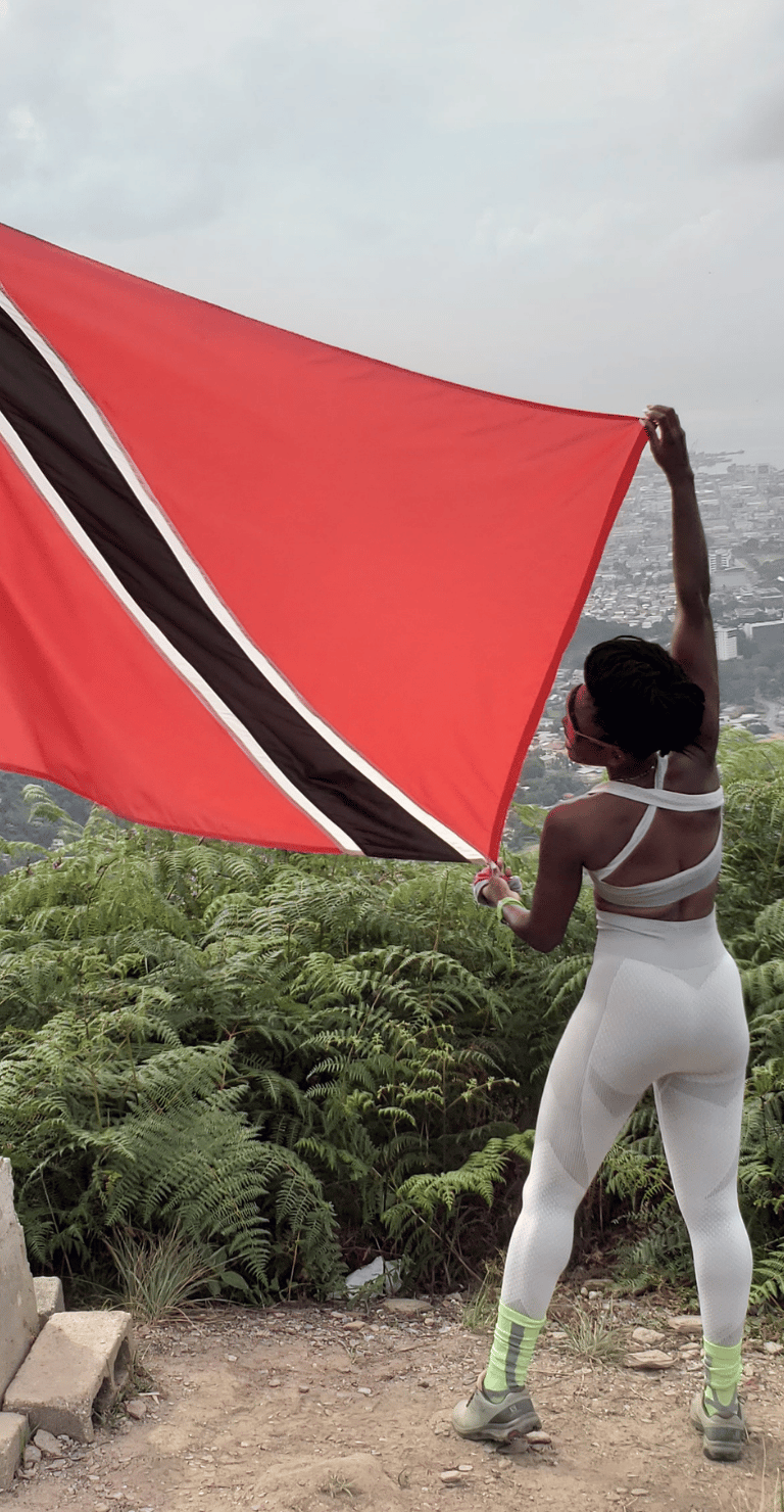 a woman holding a Trinidad and Tobago flag on top of a hill