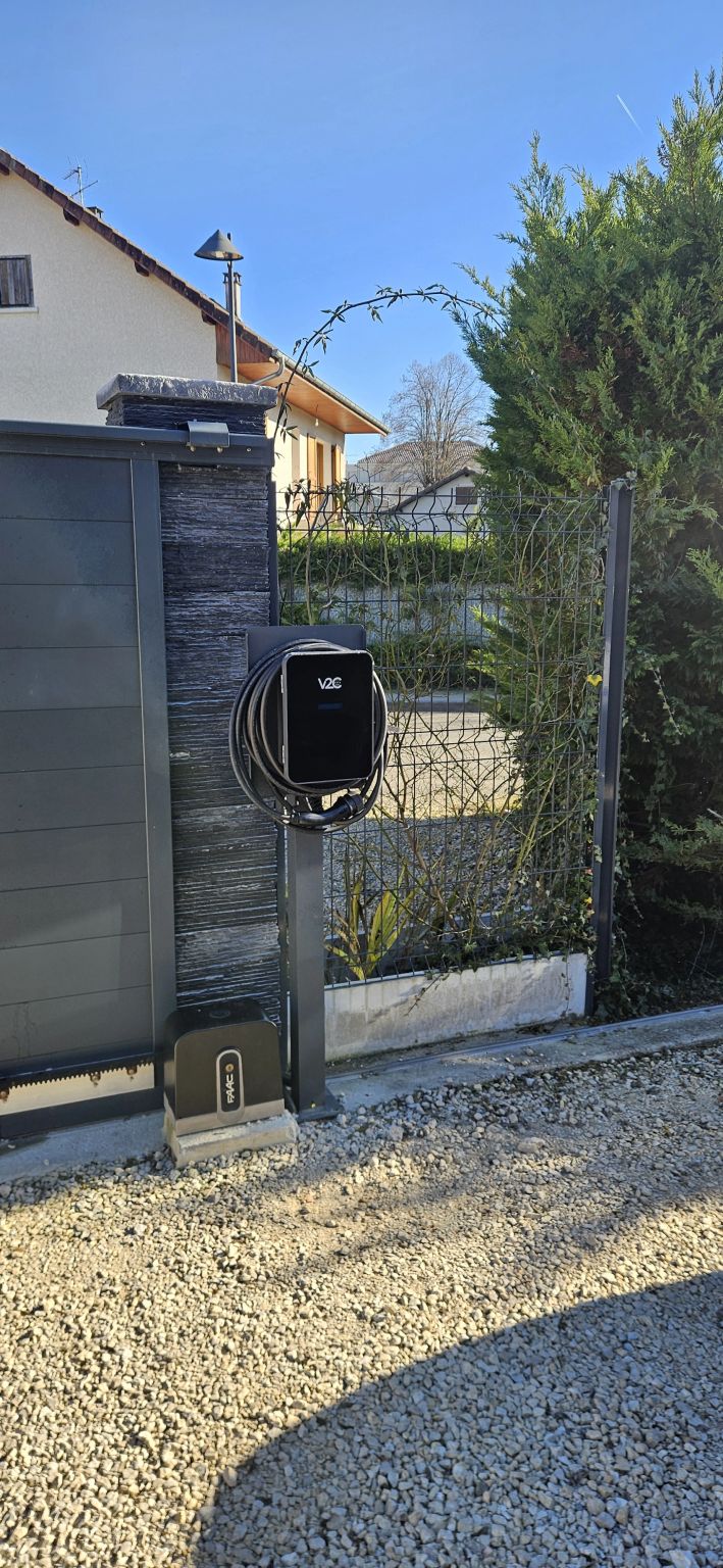 Black V2C electric vehicle wallbox charger mounted on a stone gate post in a residential driveway.