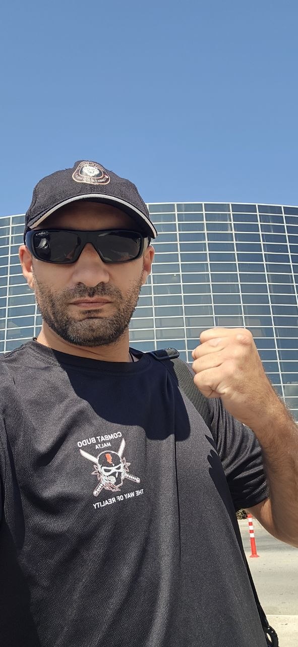 A man wearing a black martial arts shirt, sunglasses, and a baseball cap posing with a clenched fist outdoors.