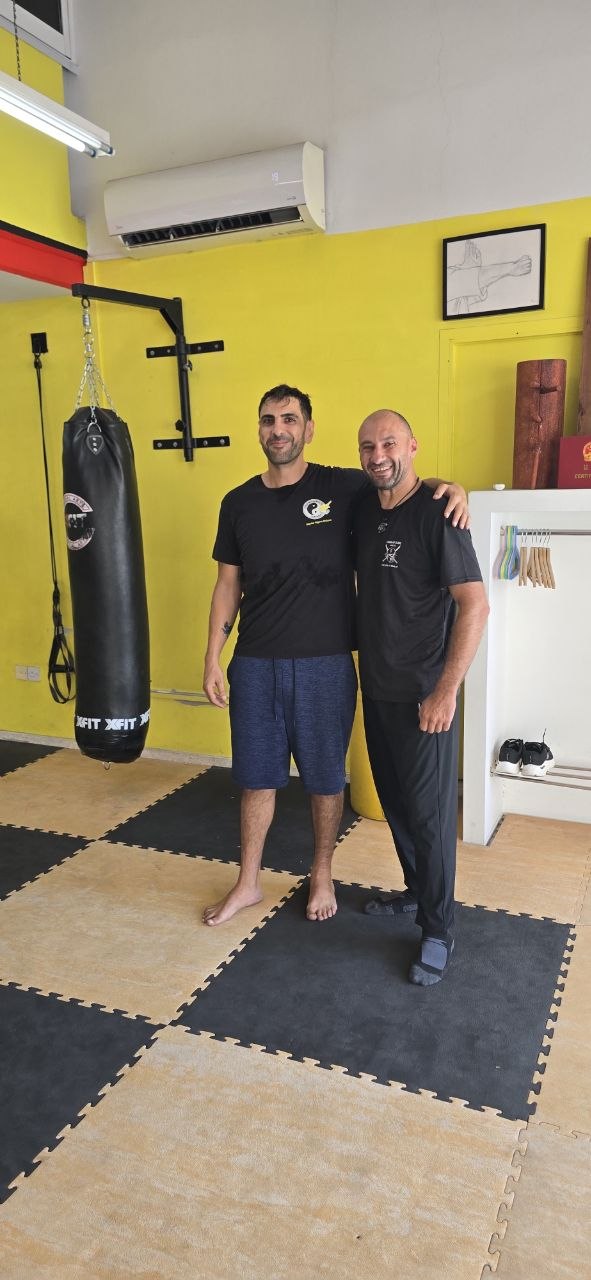 Two smiling martial arts students standing next to a heavy punching bag in a bright training gym.