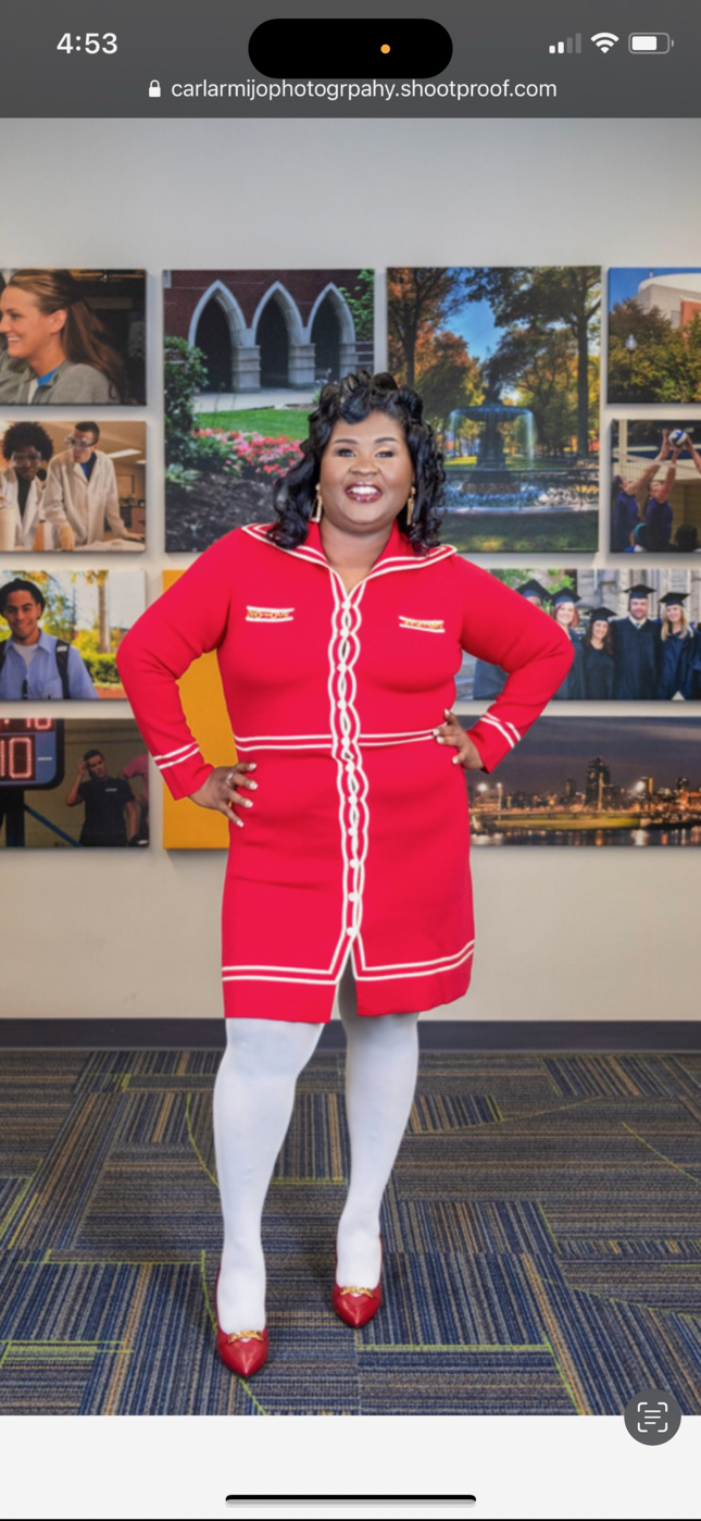 A smiling woman in a stylish red knit dress stands before a wall of campus photography.