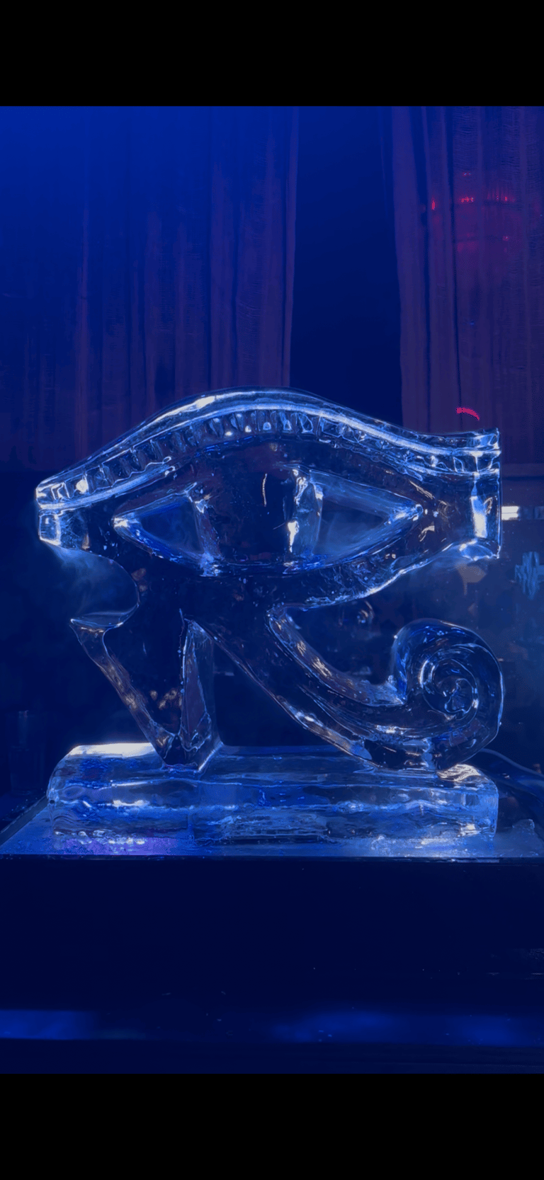 A detailed ice sculpture of the Egyptian Eye of Horus glowing with blue light in a dark room.