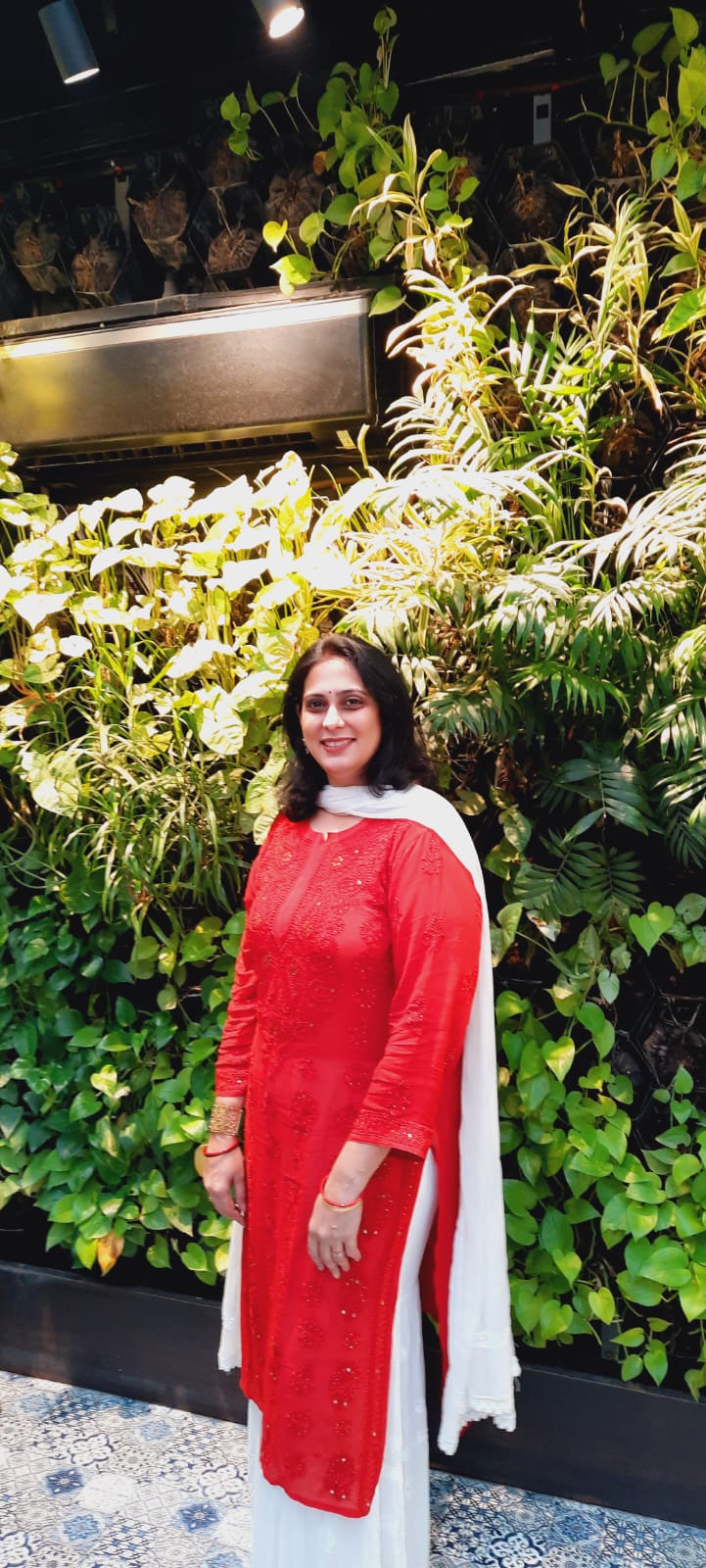 A woman in a red and white Indian ethnic suit poses against a lush vertical garden wall.