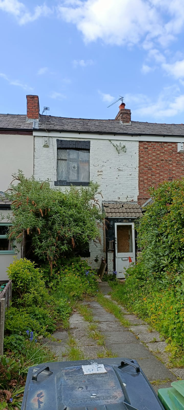 Abandoned white brick terraced house with overgrown garden and boarded windows under a blue sky.