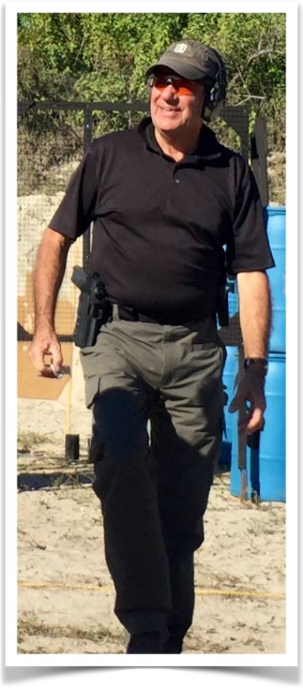 A man wearing tactical gear and a holstered handgun walks at an outdoor shooting range.