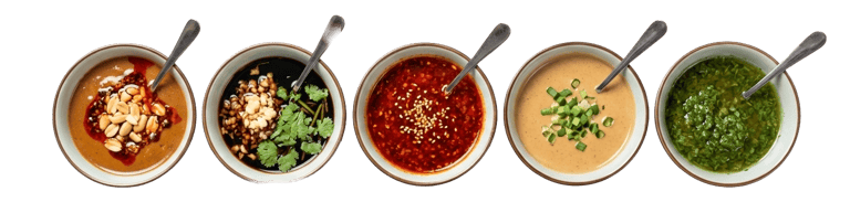 Row of five various dipping sauces in white bowls, including sesame, soy, chili oil, and fresh herbs.