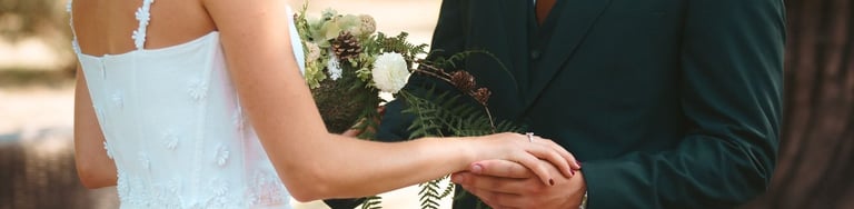 Smiling bride in white floral gown and groom in forest green suit at an outdoor wedding.