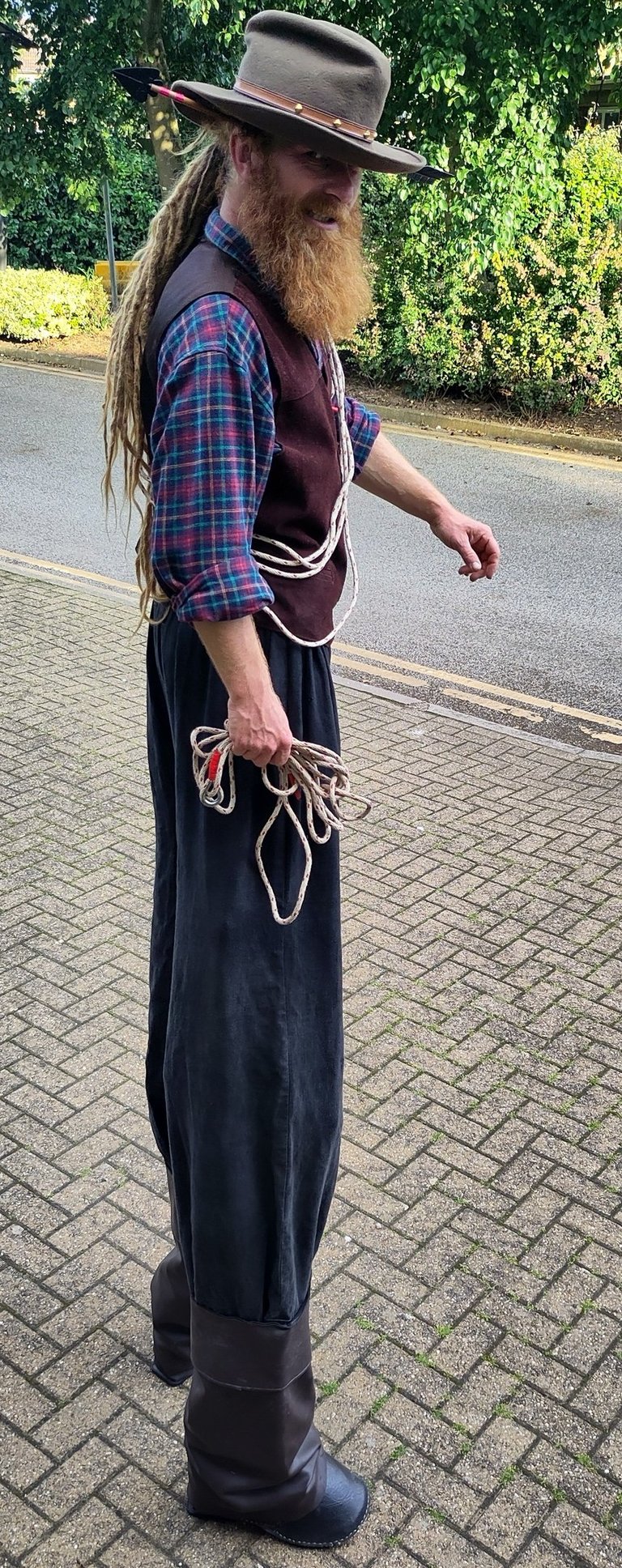 A stiltwalker with a long ginger beard wearing a cowboy hat and western attire holding a lasso