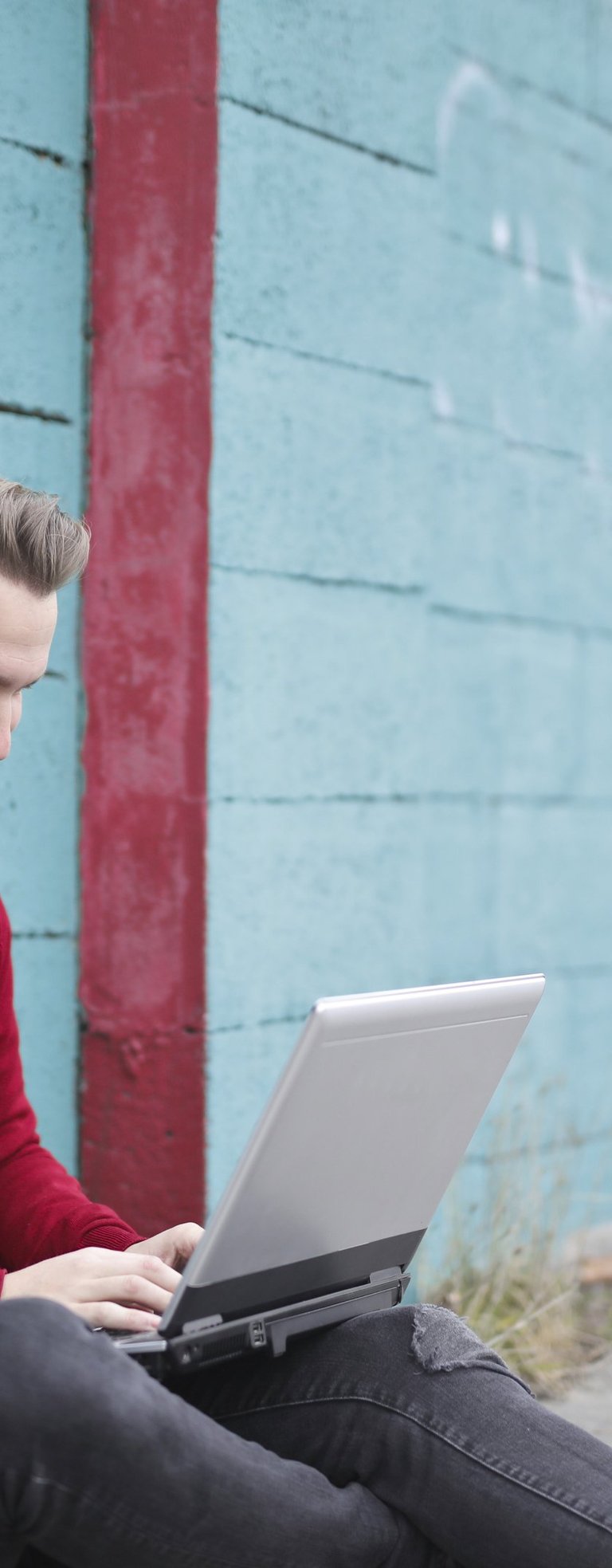 A man sits against a wall using a laptop. He is wearing a red jumper and is smiling.