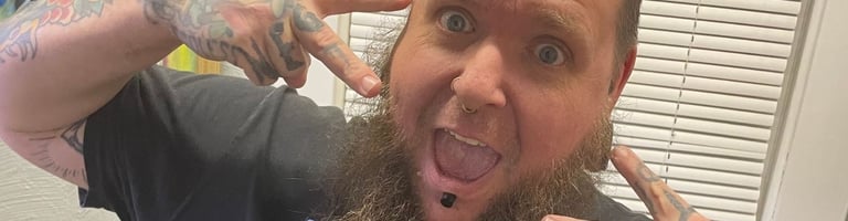 Fecke poses in his piercing room, making peace signs, an excited face, long beard, septum & lip jewelry, & tattooed hands.