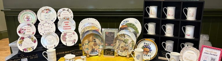 A display of Swears of Joy sassy china plates and mugs with humorous, sweary messages on a yellow tablecloth.