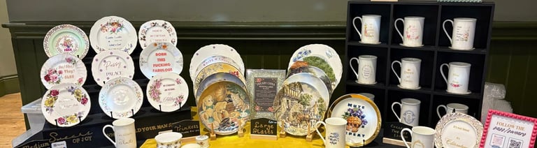 A display of Swears of Joy sassy china plates and mugs with humorous, sweary messages on a yellow tablecloth.