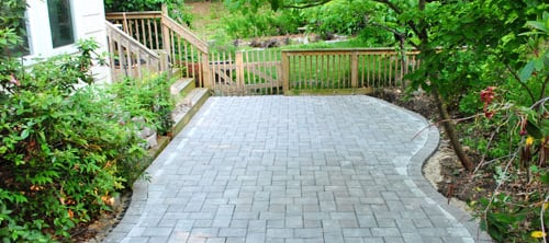 Curved grey stone paver patio installation next to a wooden deck and lush green backyard garden.