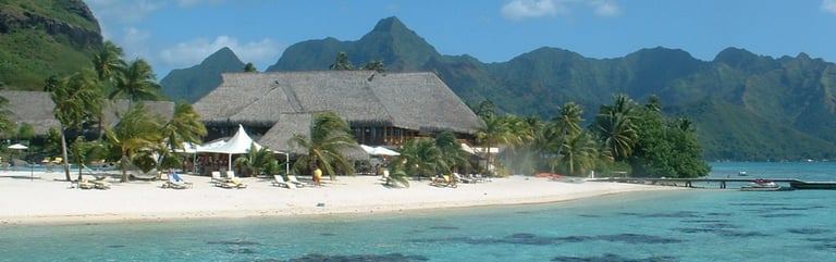 Luxury tropical resort with thatched huts on a white sand beach by turquoise ocean water and lush green mountains.