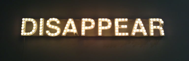 Illuminated marquee light sign spelling out DISAPPEAR in vintage bulbs against a dark wall.