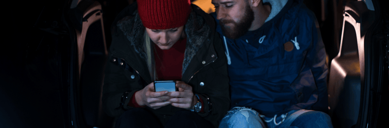 Vīrietis un sieviete sēž uz bagāžnieka vakarā un skatās telefonā