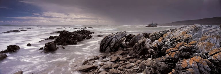 Panoramic seascape of rugged South African coastline with stormy skies and a distant shipwreck on the horizon.