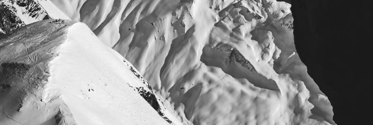 Black and white silhouette of two hikers standing on a rocky mountain ledge overlooking snowy alpine peaks.