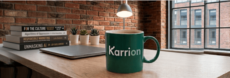 A close up of a Karrion Consulting coffee mug on a table with a laptop and books in the background.