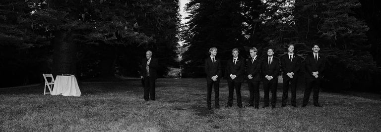 Groom and groomsmen in black suits standing in a forest for a woodland wedding ceremony.