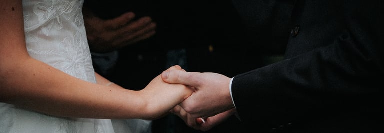 Close-up of a bride in a lace gown and groom in a black suit holding hands during a wedding ceremony.