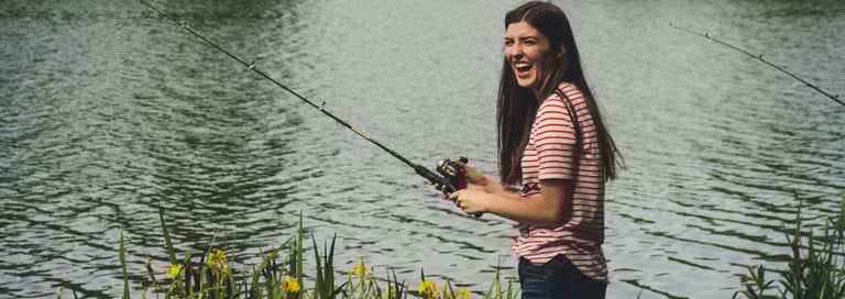 mulher morena com blusa listada pescando com vara de fibra e lago ao fundo.