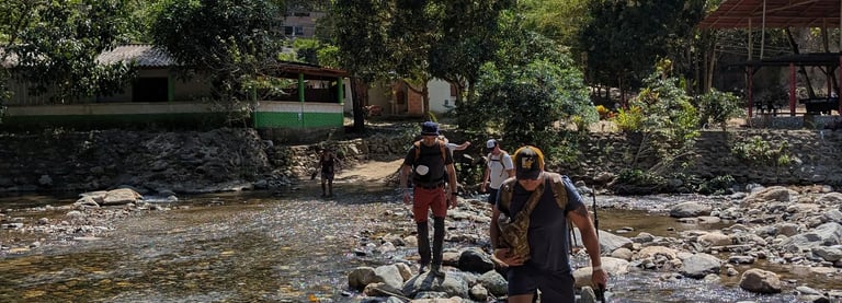 Hikers crossing a rocky river on a tropical nature trail with lush green trees and mountain views.
