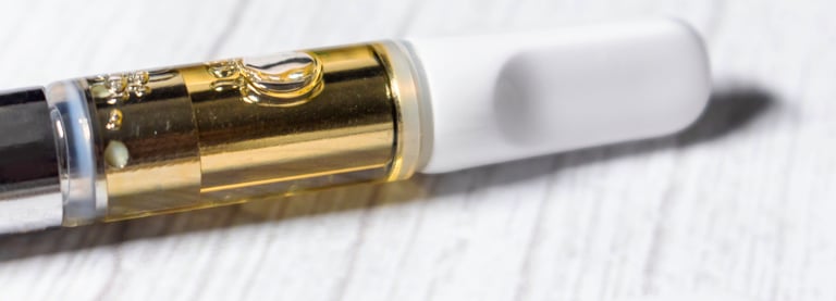 Close-up of a glass cannabis oil vape cartridge with gold distillate and white mouthpiece on a light wood surface.