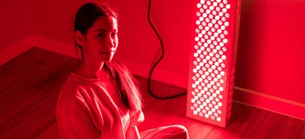A woman sits in front of a red light therapy panel for skin health and recovery.