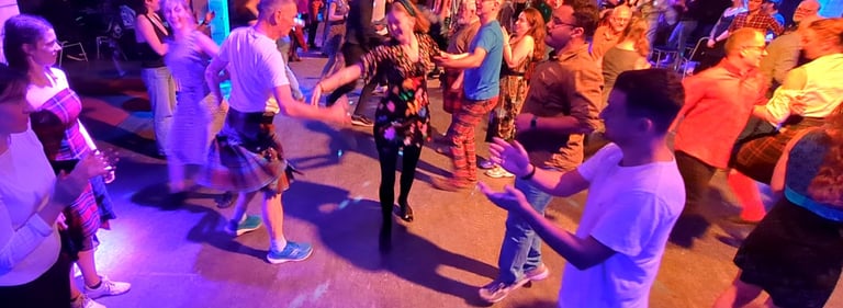 People dancing at a traditional Scottish ceilidh night with a live band and purple stage lighting.