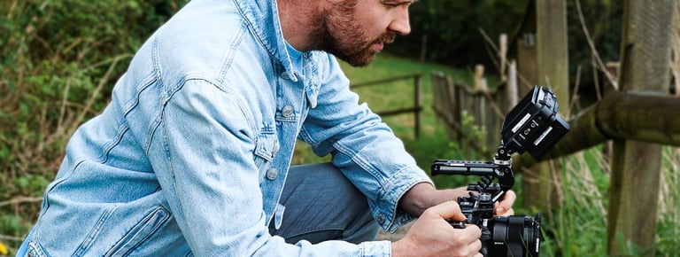Mann mit Bart in blauer Jeansjacke, draußen, hockend, mit Kamera Rig in den Händen, seitliches Profi