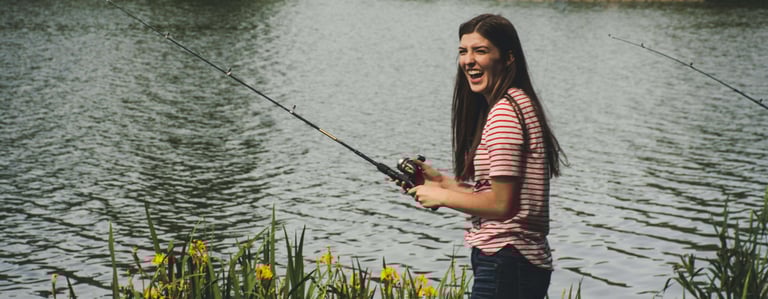 mulher morena com blusa listada pescando com vara de fibra e lago ao fundo.