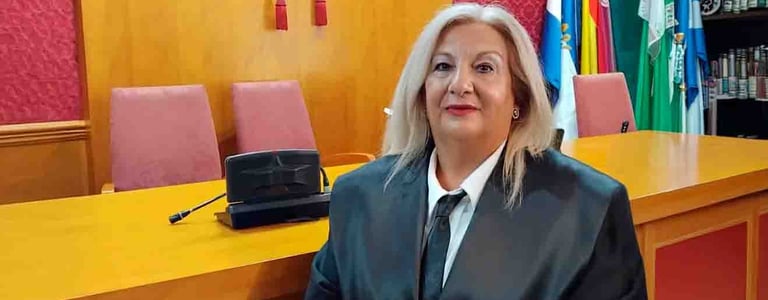 Abogada con bata negra de pie en una sala del tribunal con banderas oficiales y un emblema judicial