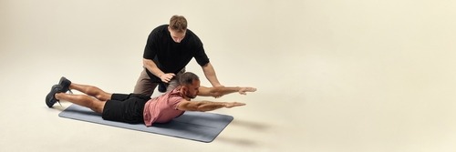 A fitness trainer assisting a man performing a back extension exercise on a yoga mat.