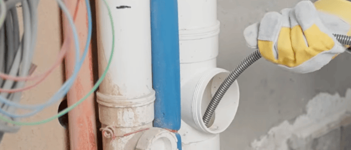 Gloved plumber's hand inserting a drain snake into a PVC pipe to clear a clog in a home's plumbing system.
