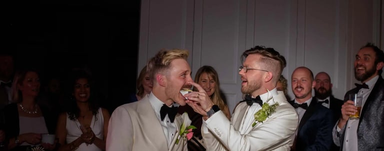 Groom in a white tuxedo feeding cake to his husband at their formal wedding reception.