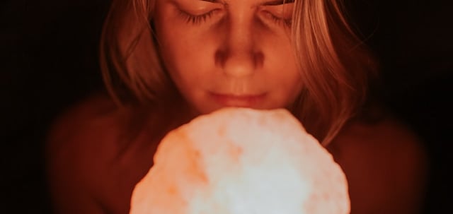 A woman with eyes closed holding a glowing pink Himalayan salt lamp in a dark room.