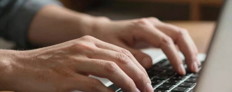 A close-up shot of hands typing on a metallic laptop keyboard, with a blurred bookshelf in the background. Natural North American daylight.