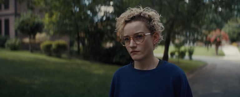 Julia Garner stands solemnly in a navy sweater and round glasses, framed by a suburban neighborhood
