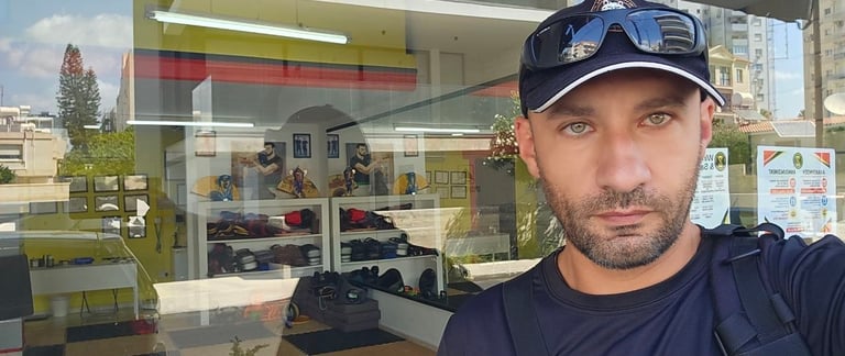 A man in a baseball cap and sunglasses takes a selfie in front of a martial arts studio window.