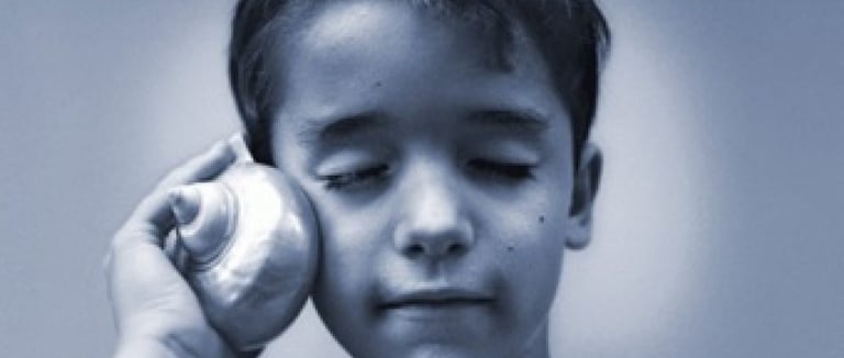 Um menino, segurando uma concha grande próxima ao ouvido, com os olhos fechados