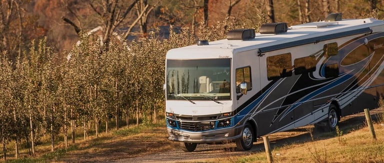A white Fleetwood Bounder Class A motorhome driving through a scenic rural apple orchard at sunset.