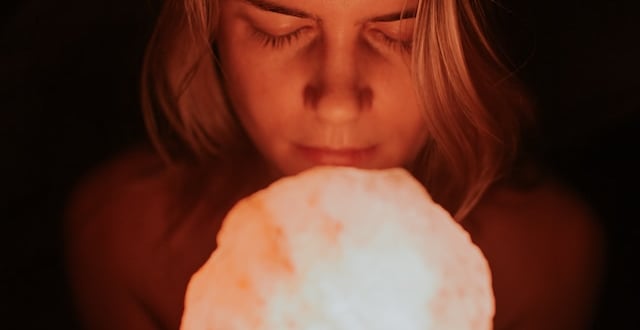 A woman with eyes closed holding a glowing pink Himalayan salt lamp in a dark room.