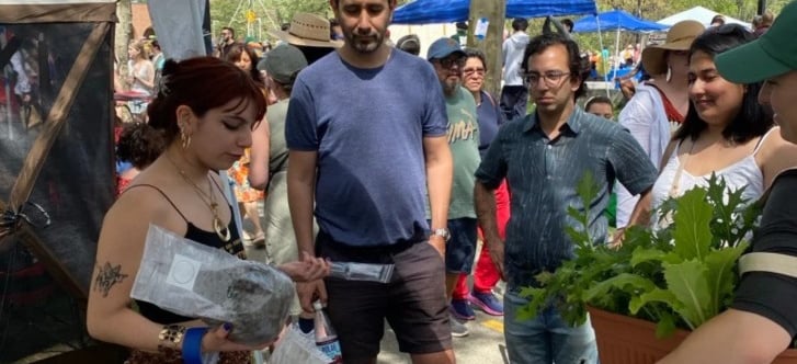 Customers shopping for organic plants and local goods at a crowded outdoor community farmers market.